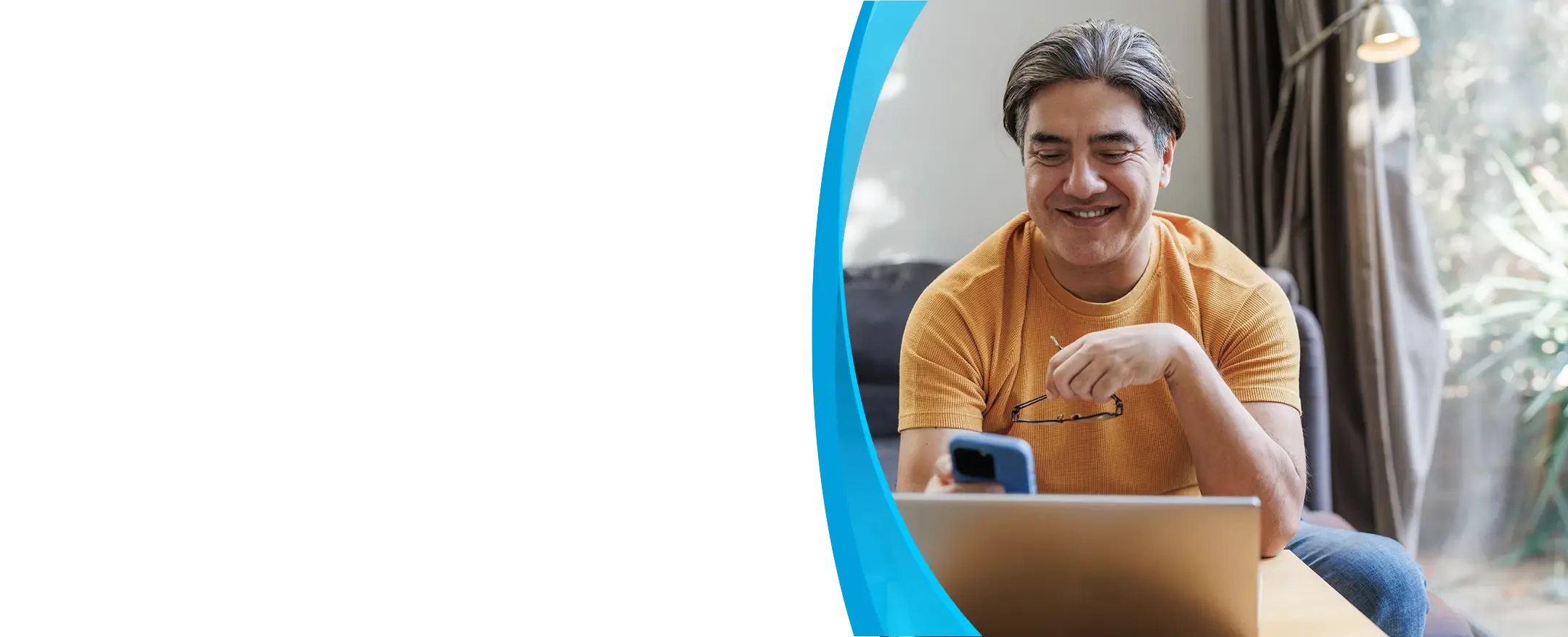 A smiling man in an orange shirt checks his phone while sitting at a laptop.